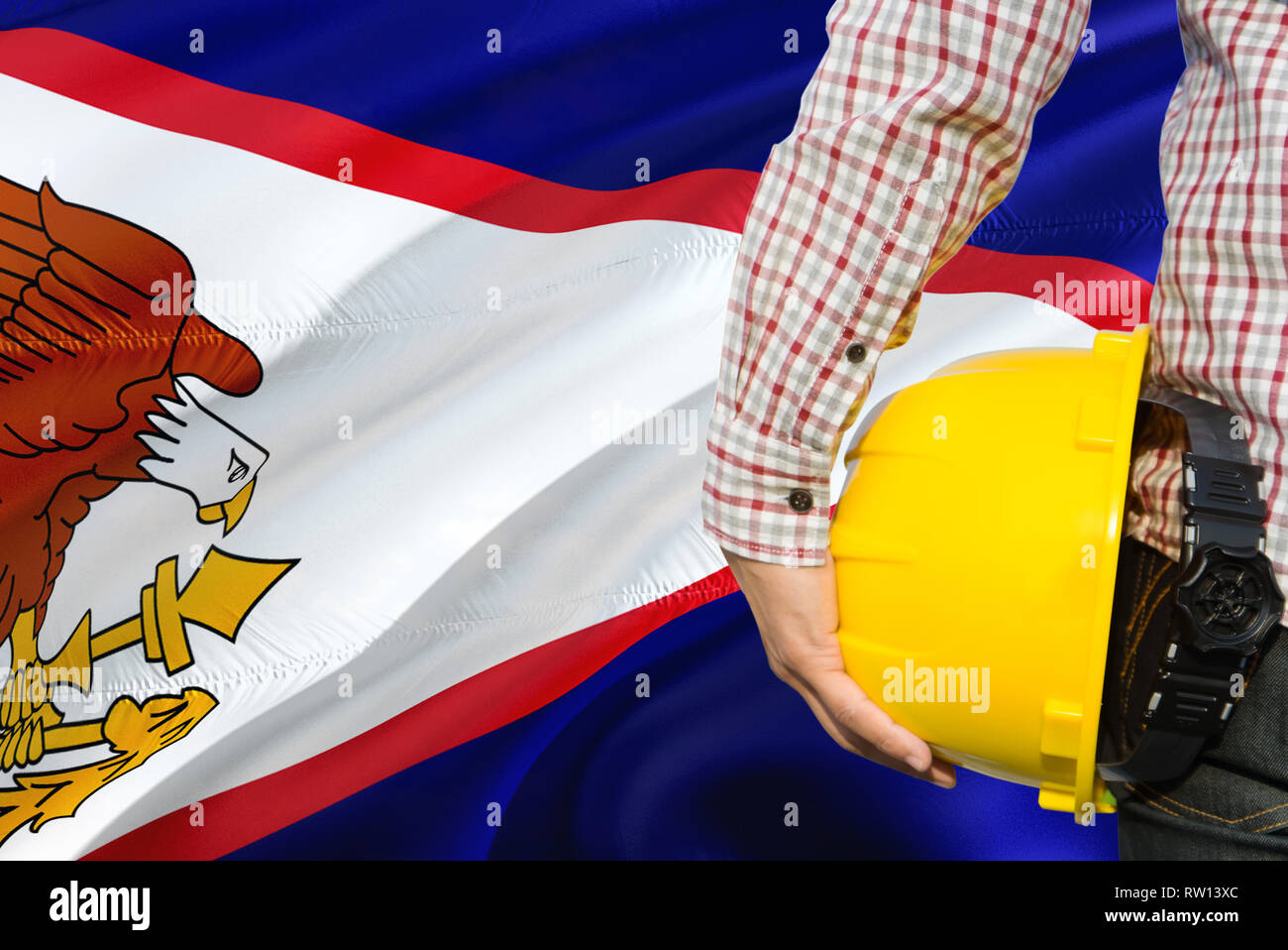 Engineer is holding yellow safety helmet with waving American Samoa flag background. Construction and building concept. Stock Photo