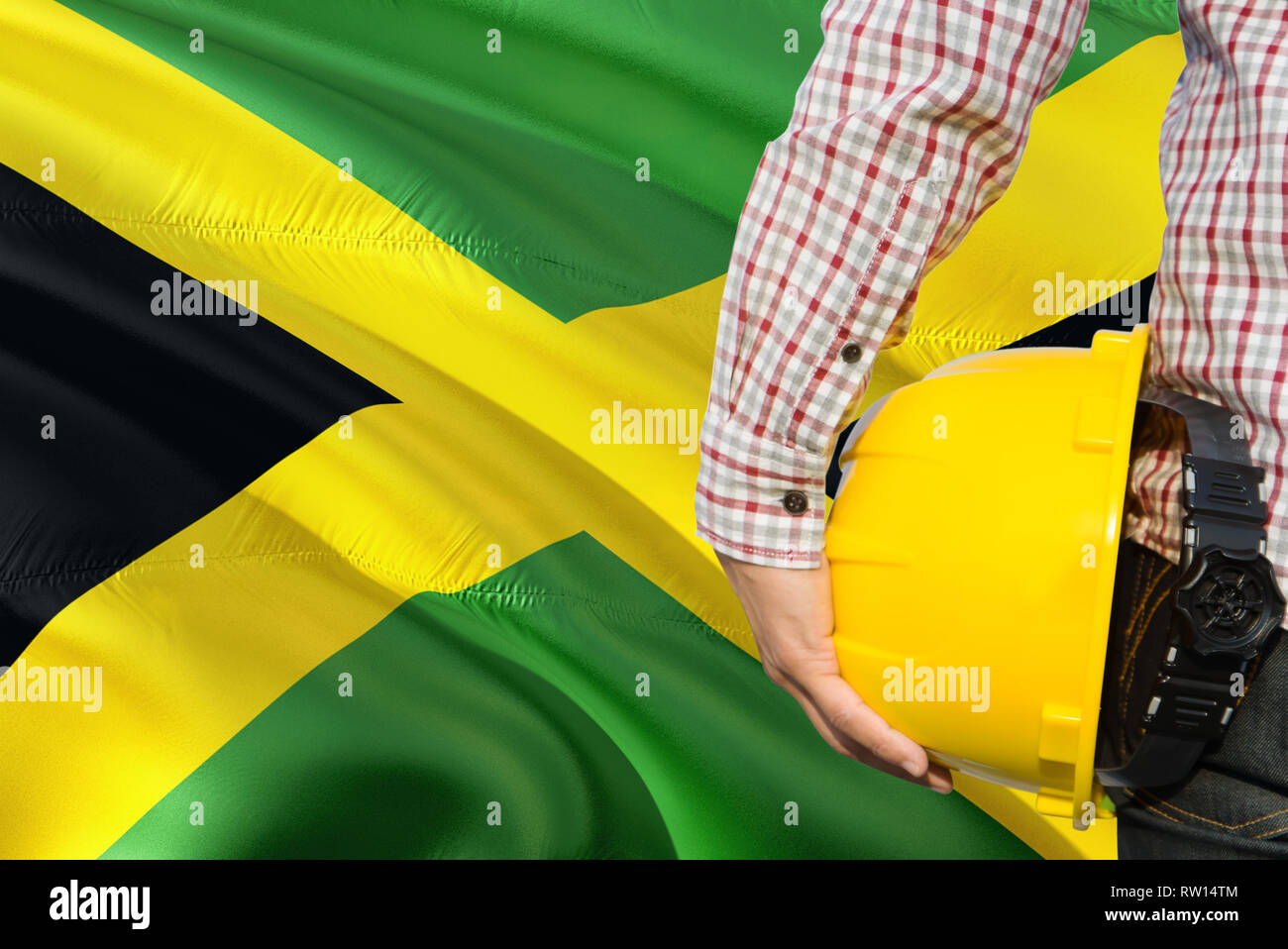 Jamaican Engineer is holding yellow safety helmet with waving Jamaica flag background