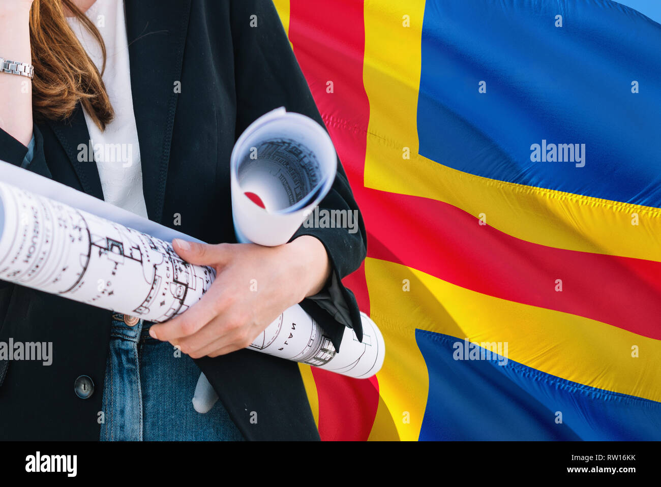 Architect woman holding blueprint against Aland Islands waving flag background. Construction and architecture concept. Stock Photo