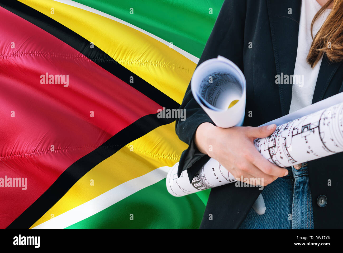 Guyanese Architect woman holding blueprint against Guyana waving flag background. Construction and architecture concept. Stock Photo