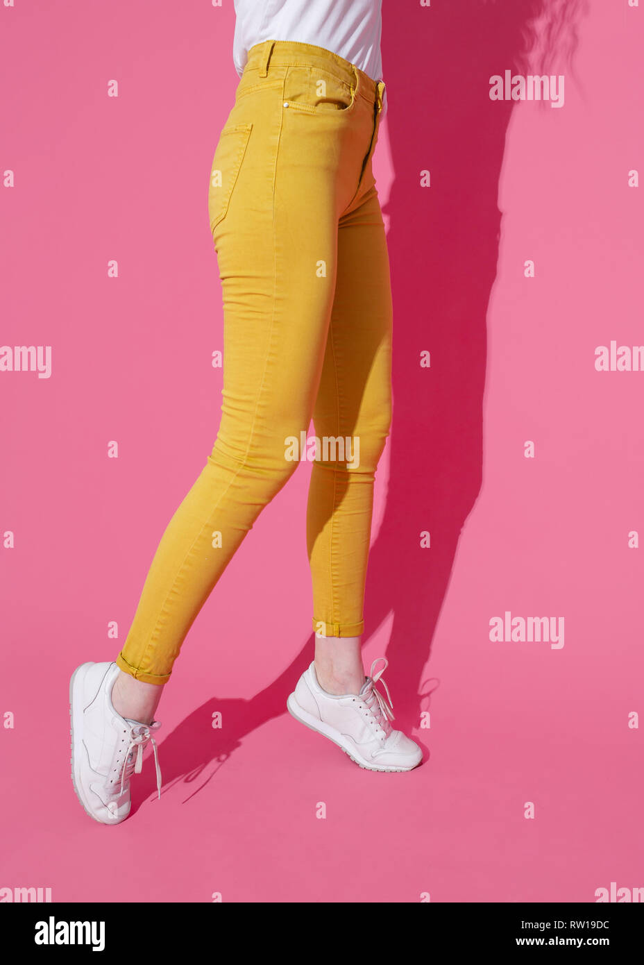 Female feet in trendy yellow jeans and white sneakers on a pink background. Stock Photo