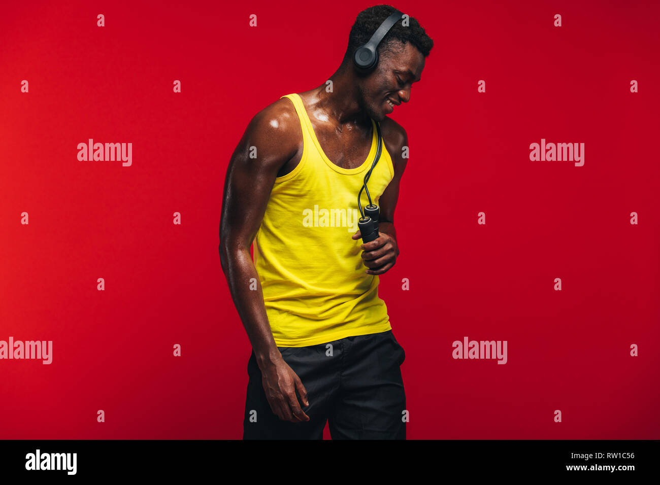 African muscular man with jumping rope listening to music on headphones on red background. Fit young man relaxing after workout. Stock Photo