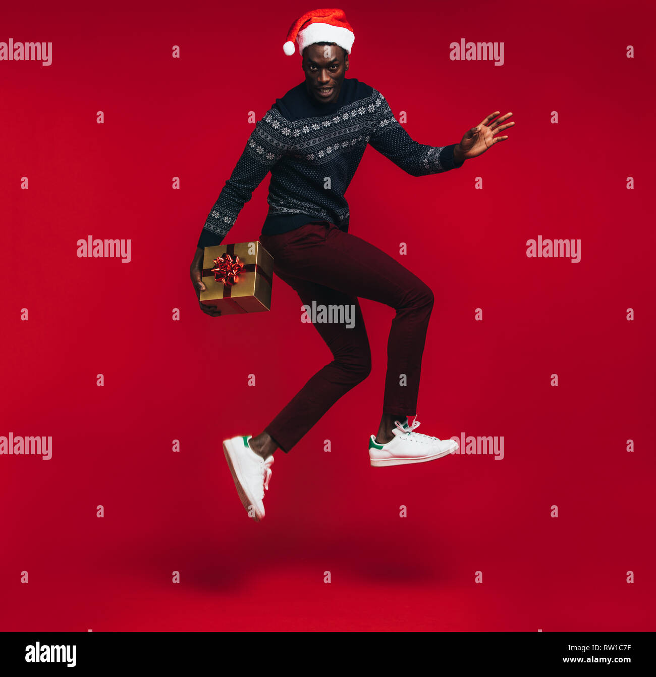 Young african man wearing santa hat jumping with gift box over red background. Stylish young guy with a christmas present jumping over red background. Stock Photo