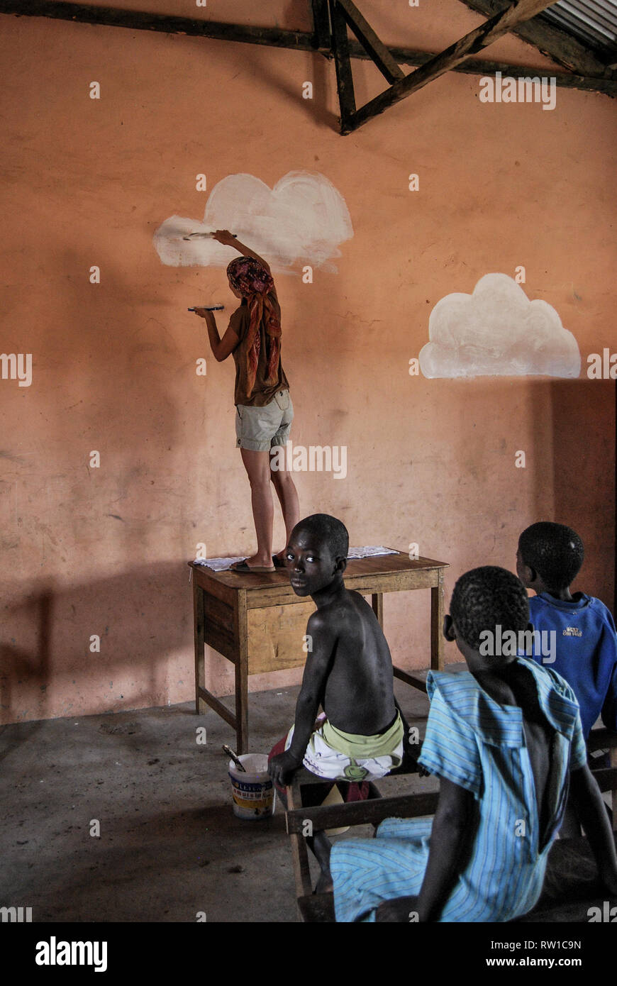 A photo of a woman painting white clouds on a wall of a local elementary school in Ghana, Africa. Students are sitting nearby and watching. Stock Photo