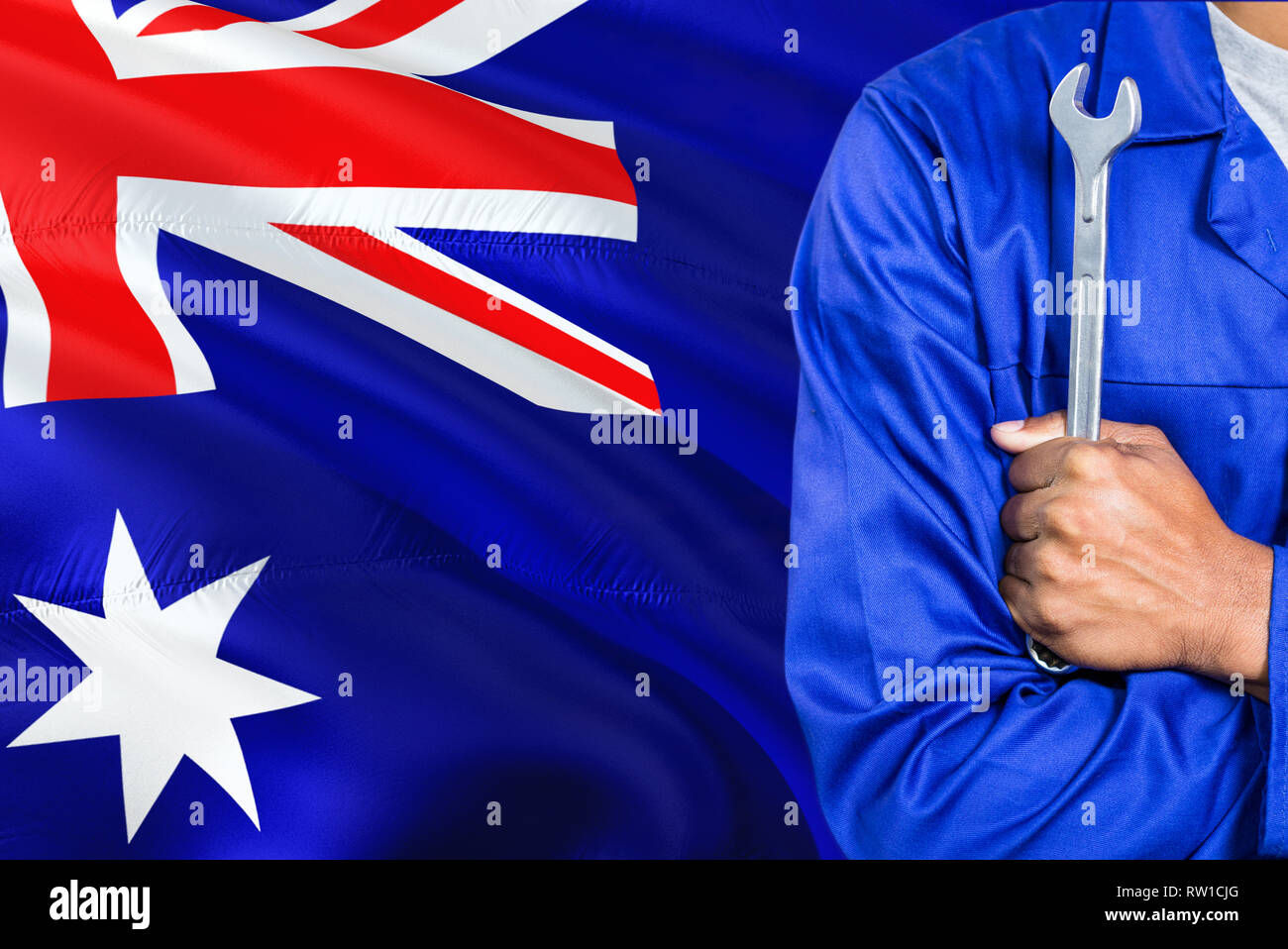 Australian Mechanic in blue uniform is holding wrench against waving Australia flag background. Crossed arms technician. Stock Photo