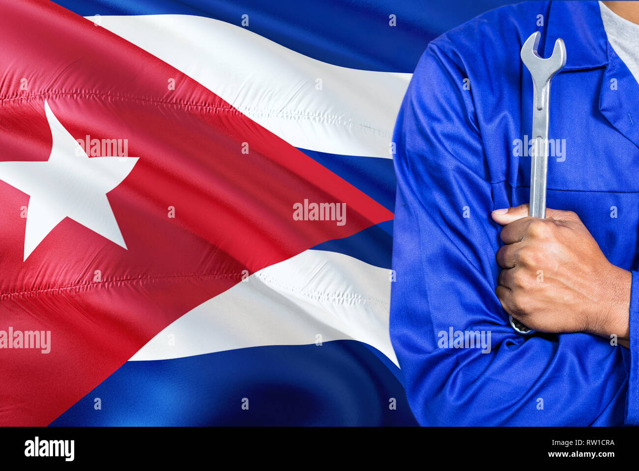 Cuban Mechanic in blue uniform is holding wrench against waving Cuba flag background. Crossed arms technician. Stock Photo