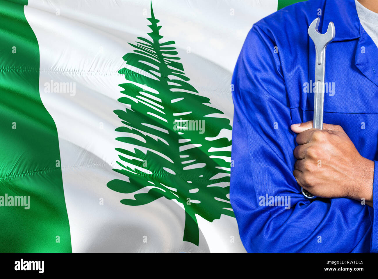 Mechanic in blue uniform is holding wrench against waving Norfolk Island flag background. Crossed arms technician. Stock Photo