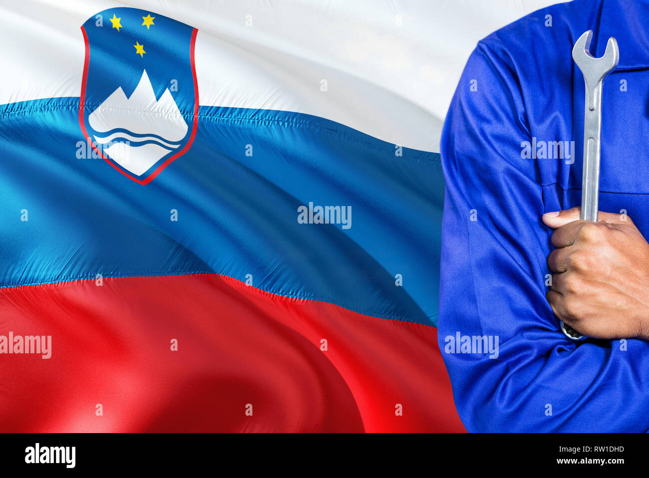 Slovenian Mechanic in blue uniform is holding wrench against waving Slovenia flag background. Crossed arms technician. Stock Photo