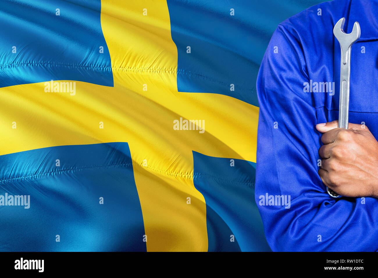 Swedish Mechanic in blue uniform is holding wrench against waving Sweden flag background. Crossed arms technician. Stock Photo