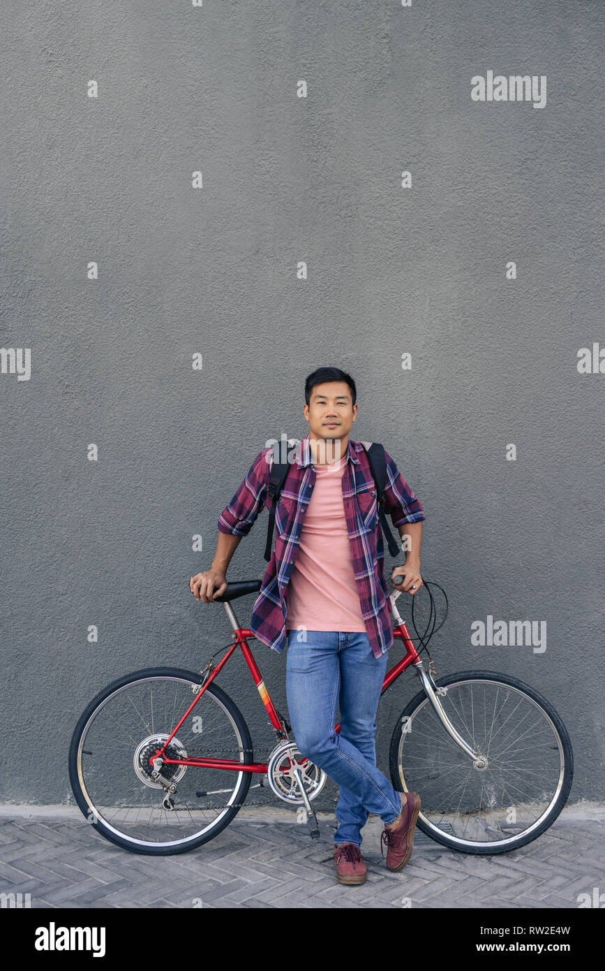 Young man standing with his bike against a gray wall Stock Photo