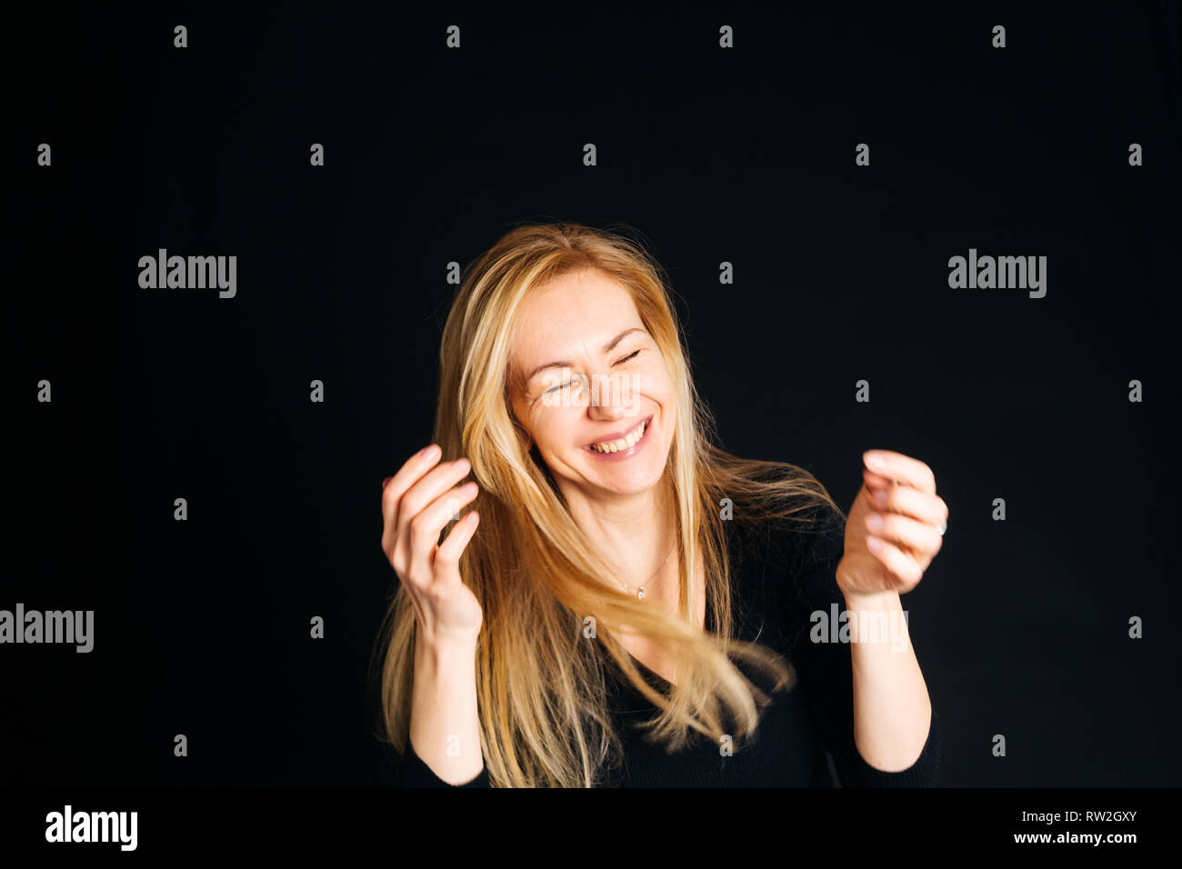 Close up studio portrait of a beautiful woman in the black dress, laughing, eyes closed, against the black background Stock Photo