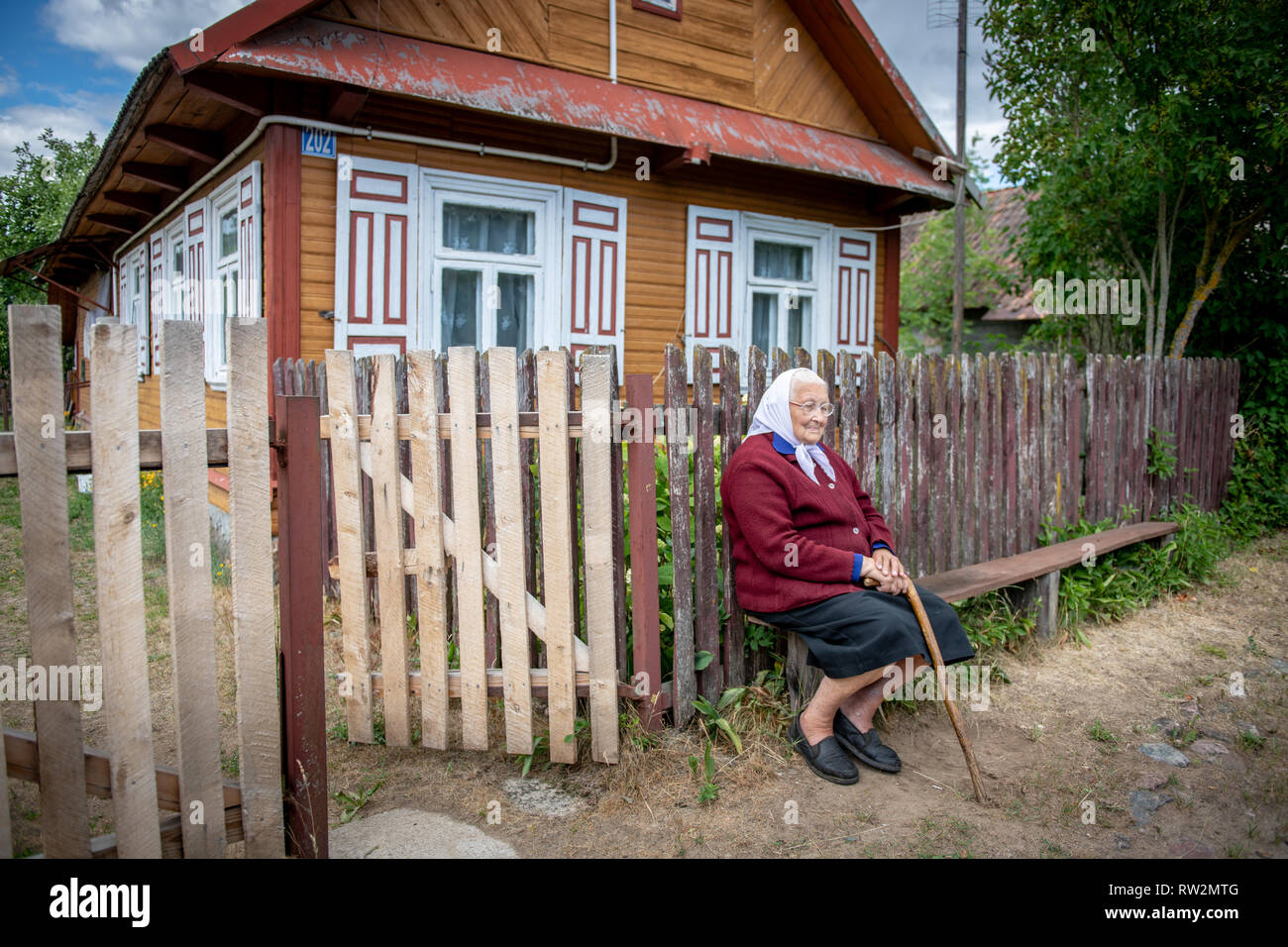 Elderly woman wearing head scarf sits on bench with hands crossed over cane in front of cabin-style home with decorative shutters in Trześcianka the ' Stock Photo