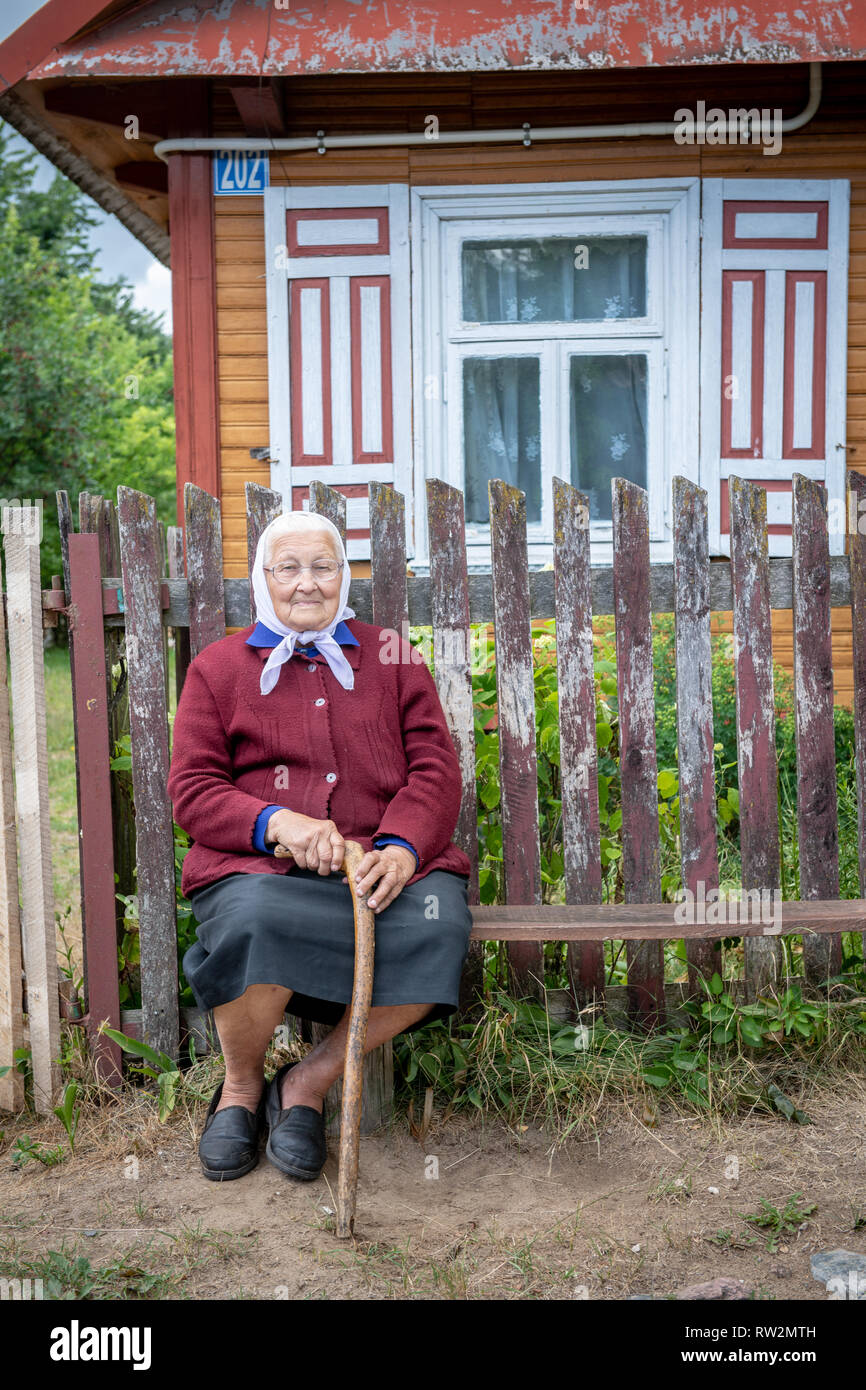 Elderly woman wearing head scarf sits on bench with cane in front of cabin-style home with decorative shutters in Trześcianka the 'Land of the Open Sh Stock Photo
