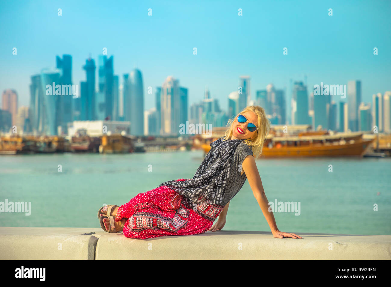 Happy caucasian woman relaxing along Corniche promenade with Dhow Harbour and Doha West Bay skyline on background. Lifestyle tourist enjoys in Doha Stock Photo