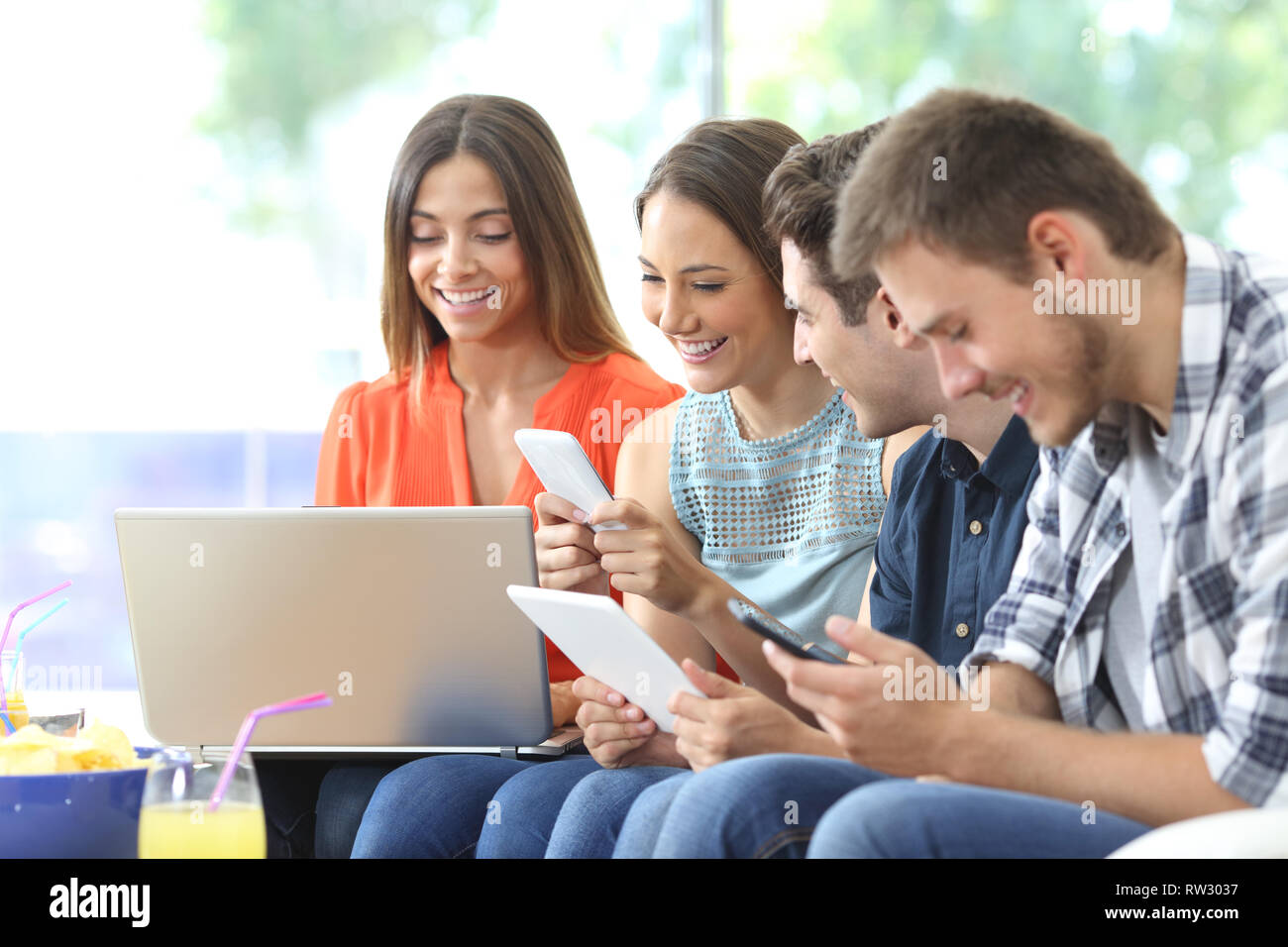 Group of happy friends using multiple devices sitting on a couch in the ...
