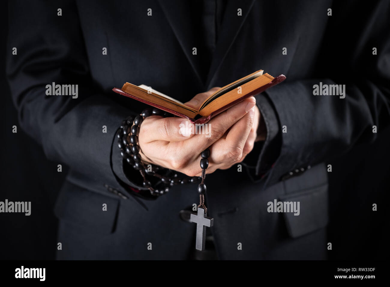 Hands of a christian priest dressed in black holding a crucifix and reading New Testament book. Religious person studies Bible and holds prayer beads, Stock Photo