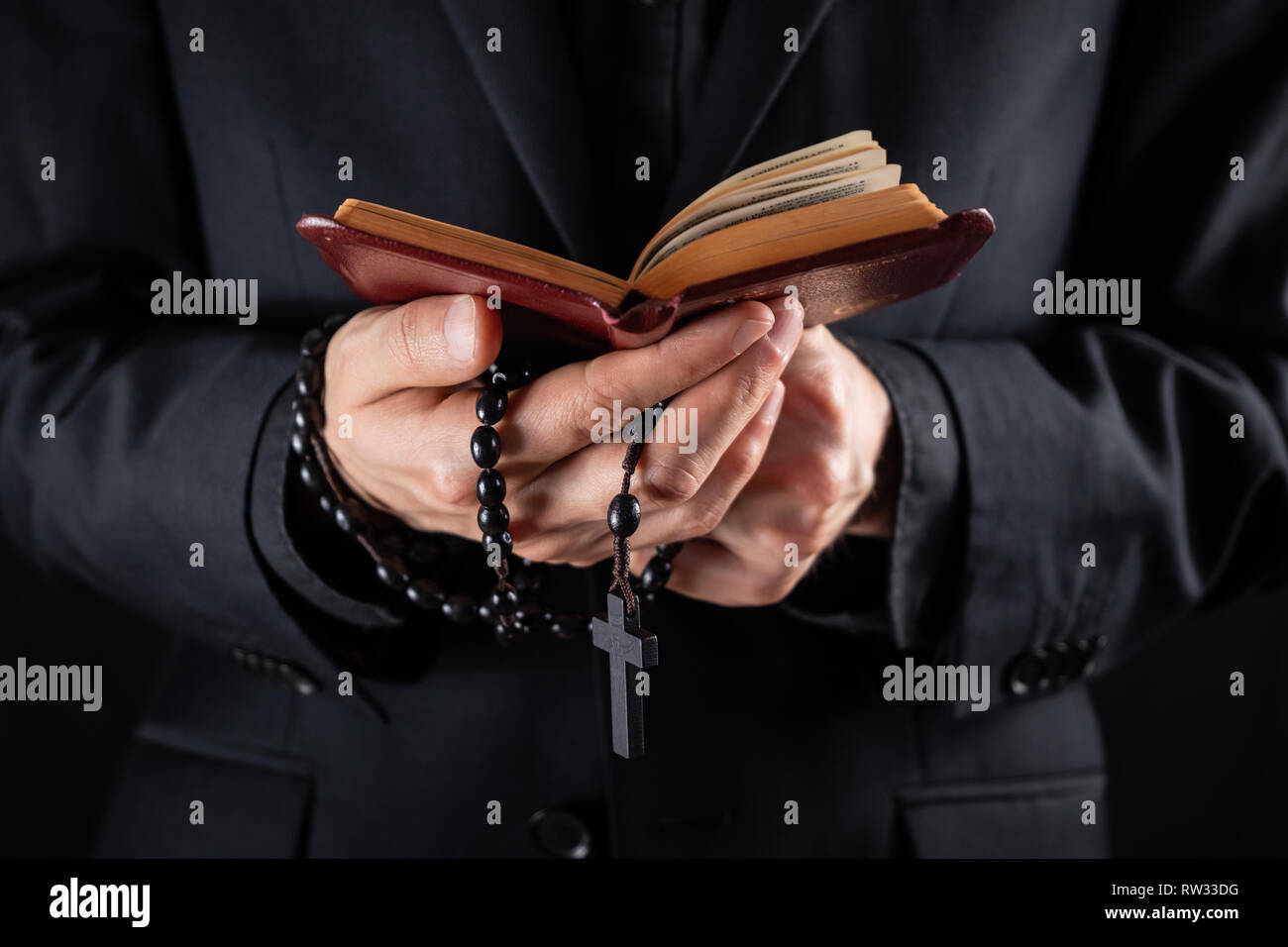 Hands of a christian priest dressed in black holding a crucifix and reading New Testament book. Religious person studies Bible and holds prayer beads, Stock Photo