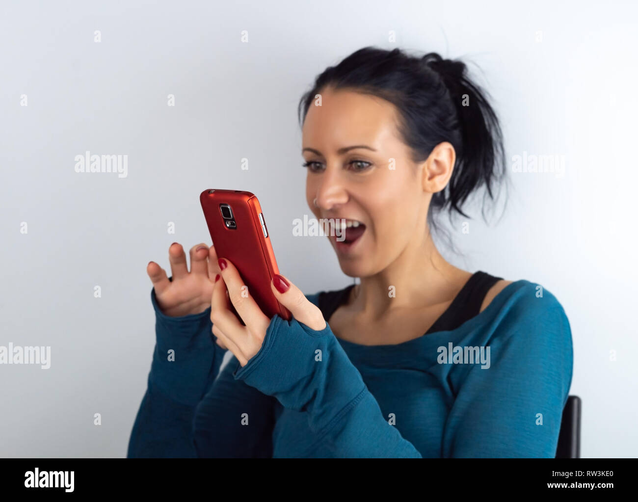 Young casual hipster woman holding and looking at cell phone reading unexpected sms news with a surprised and amazed facial expression, on white Stock Photo