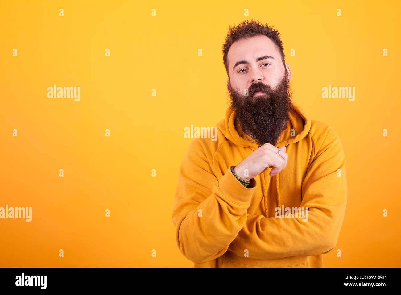 Stylish beaded hipster looking serios at the camera and playing with his beard over yellow background, Yellow hoodie. Cool man. Trendy adult. Stock Photo
