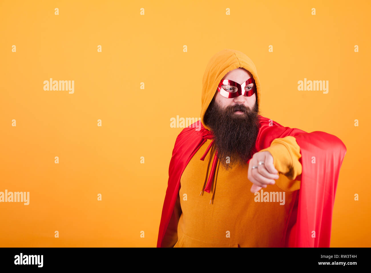 Hipster with long beard dressed in superhero costume looking away over yellow background. Yellow hoodie. red mask. Brave man. Stock Photo