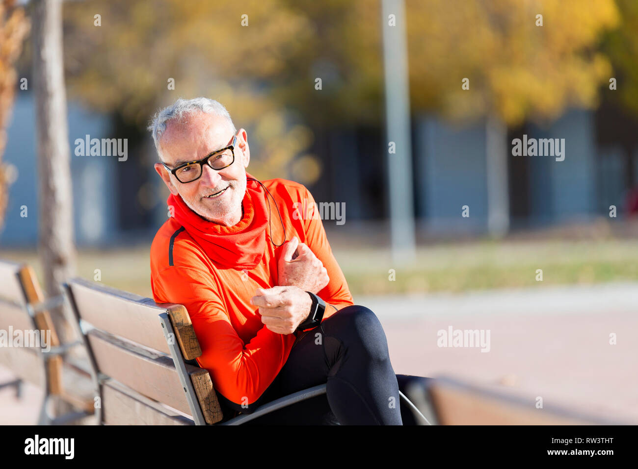 Front view of senior man in sports clothes sitting in a bench at park while smiling and looking camera in a sunny day Stock Photo