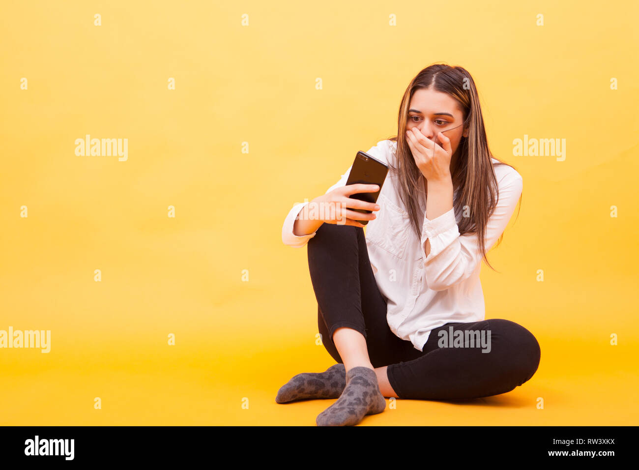 Girl looking shocked at her phone while sitting down over yellow background. Facial expression Stock Photo