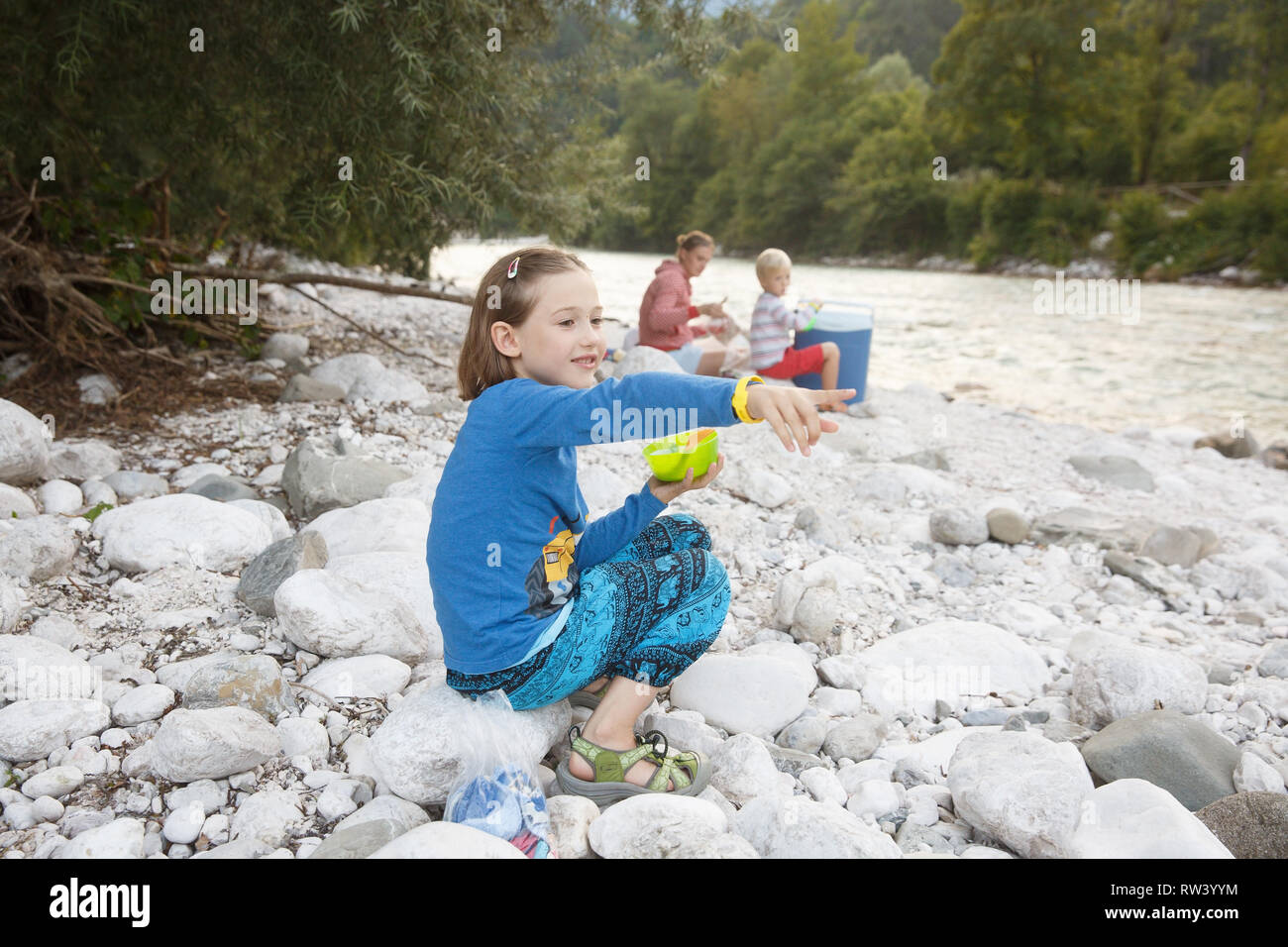 Girl eating in nature, looking at the river, having picnic with her family, sitting on the rocks on the river bank. Outdoor lifestyle, positive parent Stock Photo