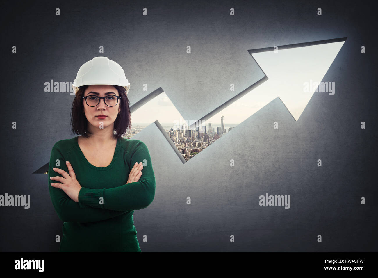 Woman engineer, wearing protective helmet, holding arms crossed, thoughtful looking over a rising graph arrow shape as a hole in concrete wall with a  Stock Photo