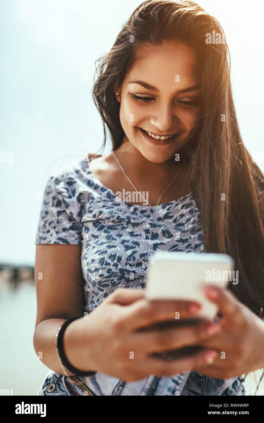 Cute smiling young woman is surfing on internet or typing message on smartphone and sharing great news with her friends. Stock Photo