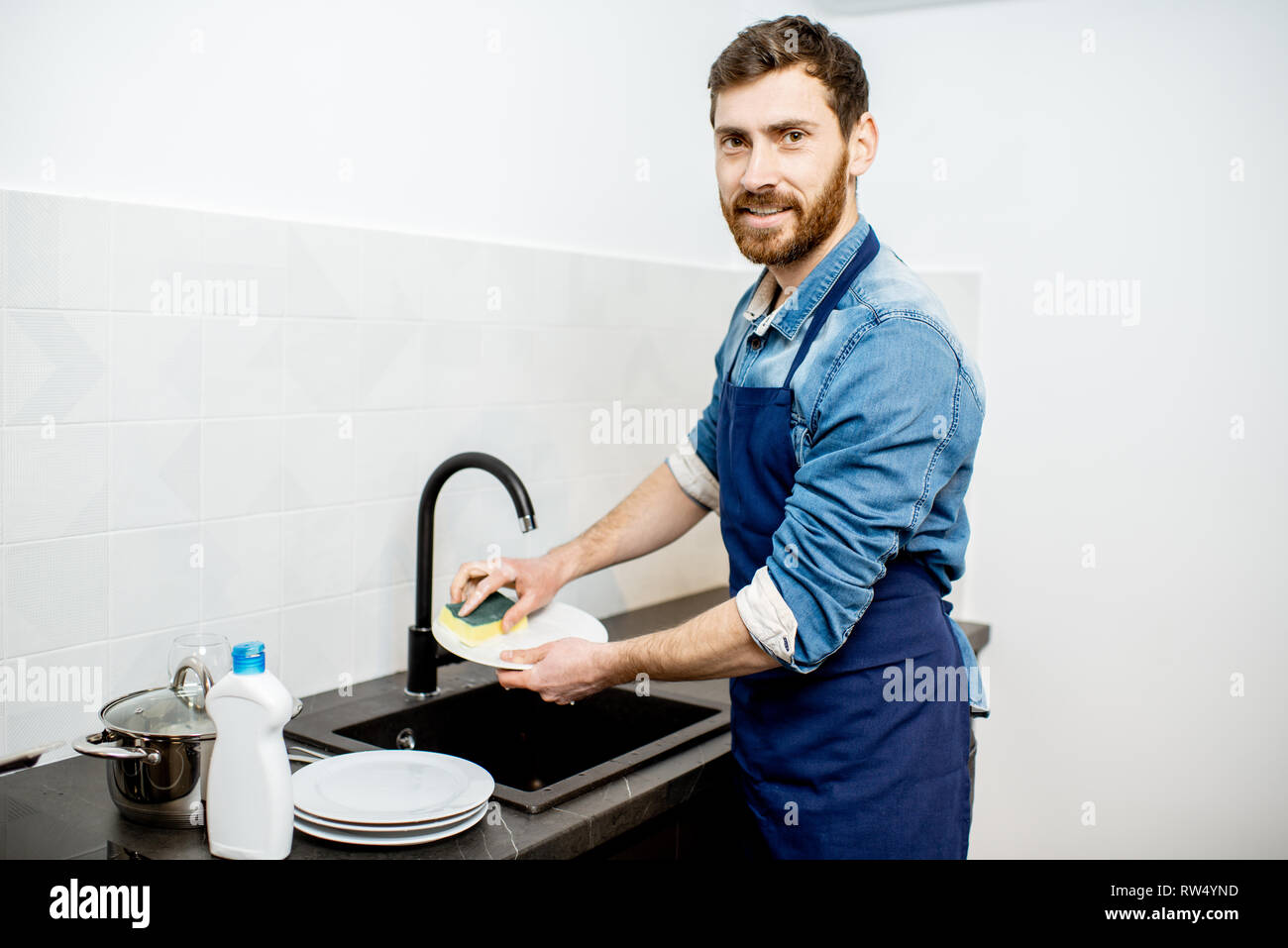 Handsome man in apron doing household chores washing dishes on the kitchen at home Stock Photo