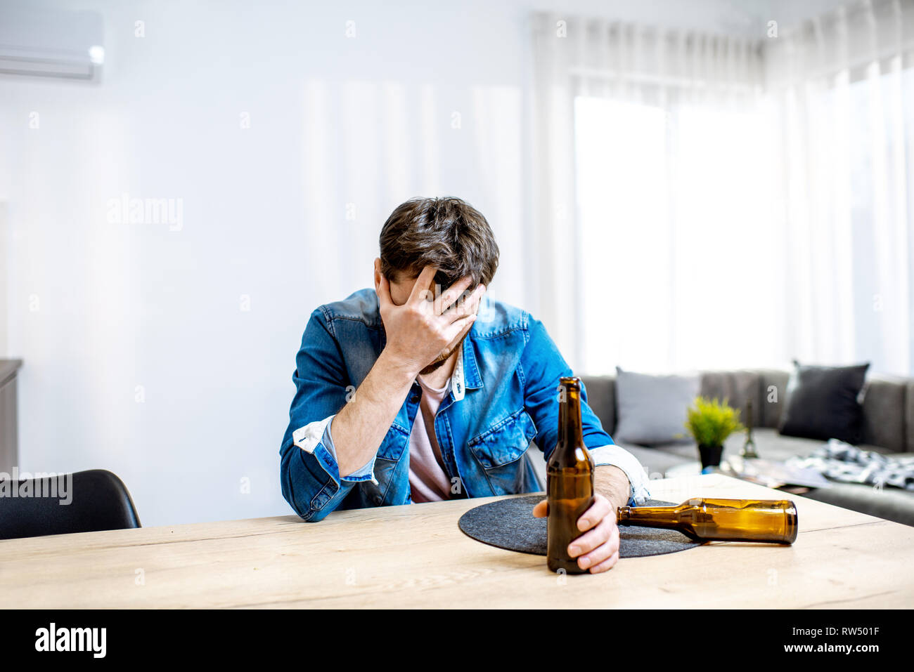 Drunk male alcoholic sitting with bottles feeling depressed and suffering from head ache at home Stock Photo