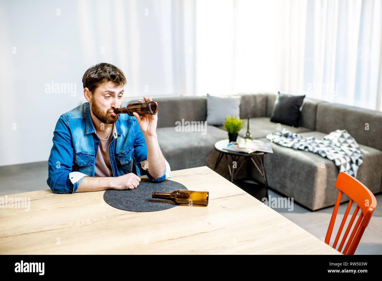 Drunk male alcoholic sitting with bottles feeling depressed and suffering from head ache at home Stock Photo