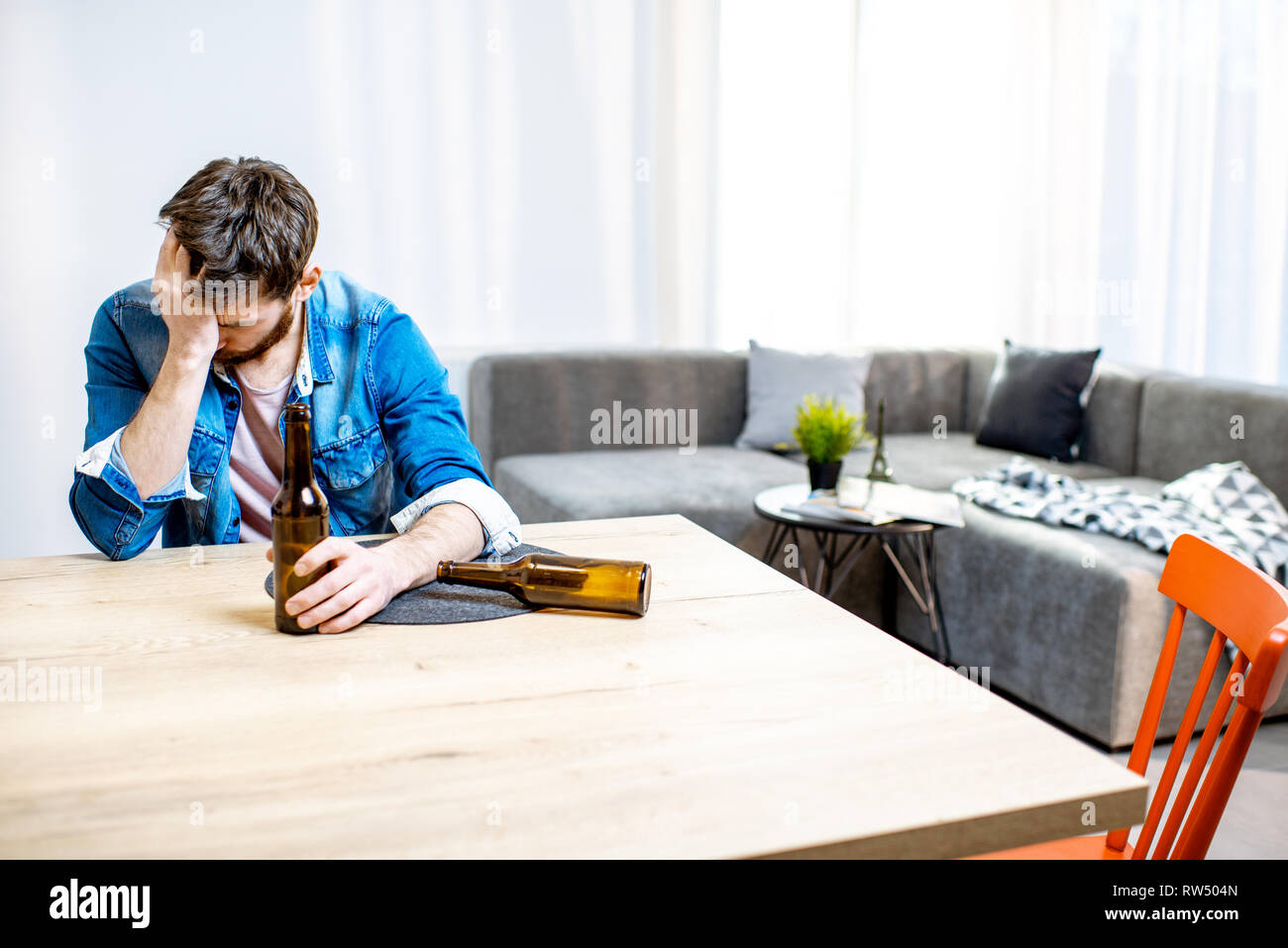 Drunk male alcoholic sitting with bottles feeling depressed and suffering from head ache at home Stock Photo