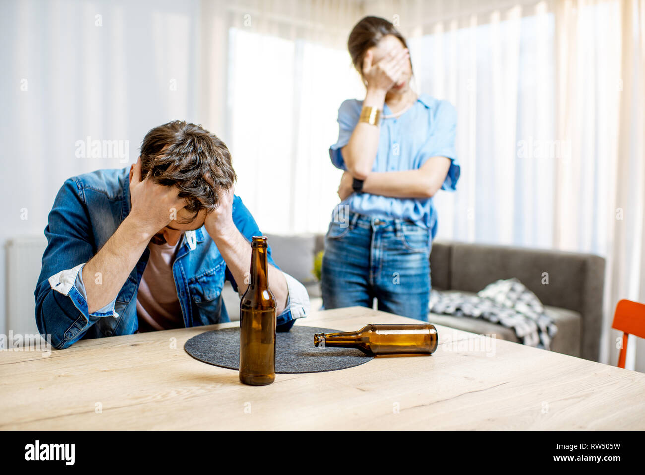 Drunk man suffering from alcoholism feeling depressed sitting at home with young woman in despair on the background Stock Photo