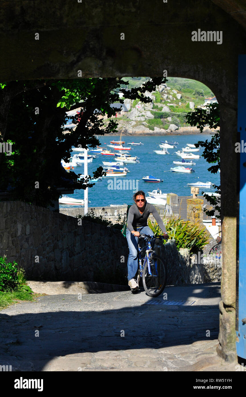 A young lady cyclist arrives at the top of the steep hill to the Garrison gate with a view of the sunlite harbour behind her. St Marys, Isles of Scill Stock Photo