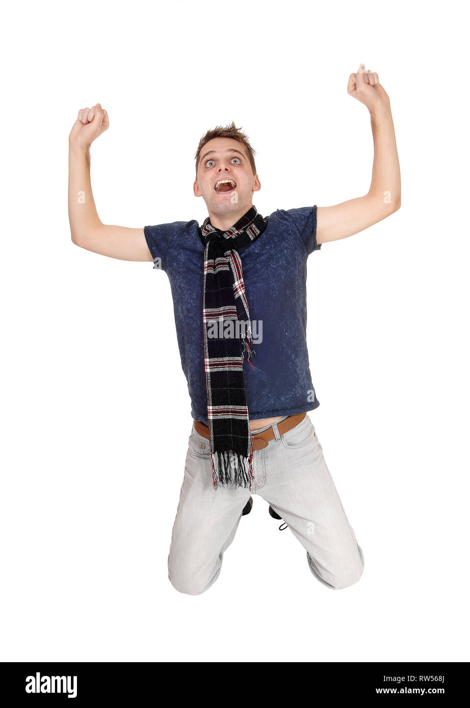 A Joyful happy young man on his knees with his arm up into the air screaming, wearing jeans and a scarf, isolated for white background Stock Photo