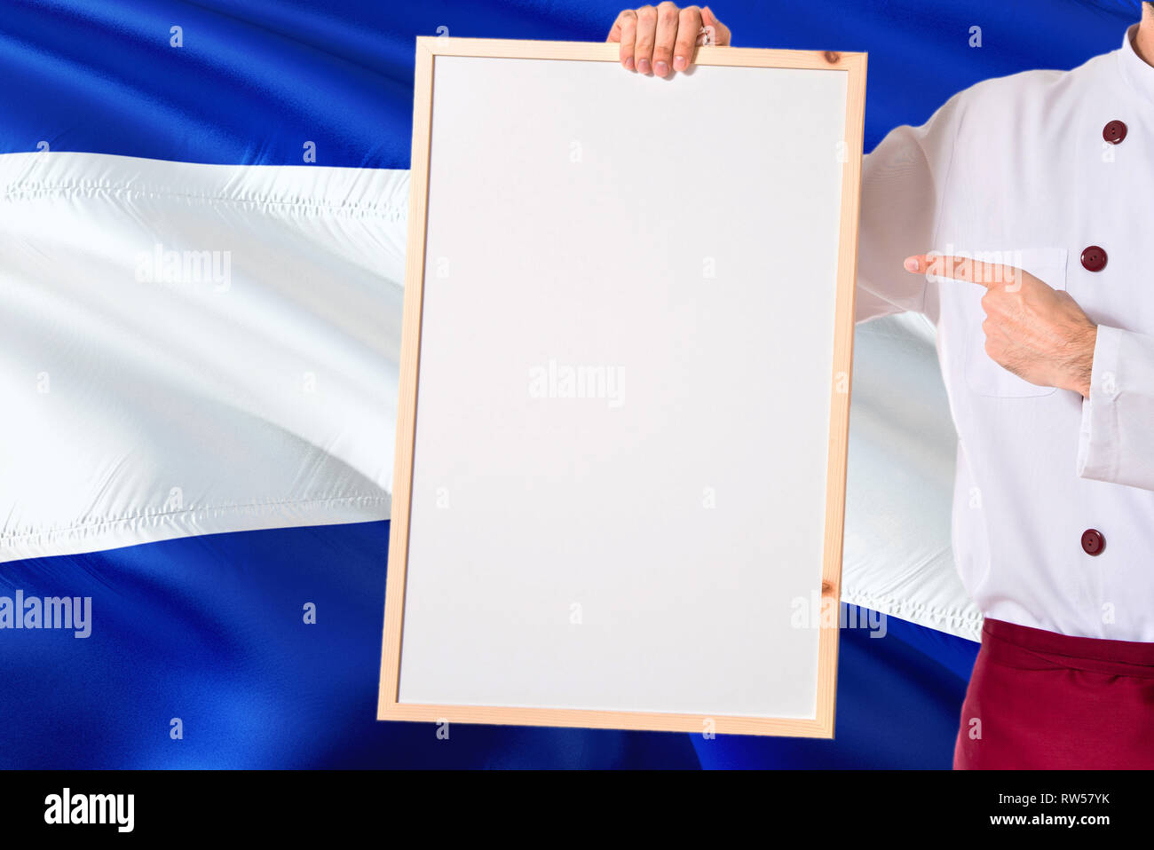 Salvadoran Chef holding blank whiteboard menu on El Salvador flag background. Cook wearing uniform pointing space for text. Stock Photo
