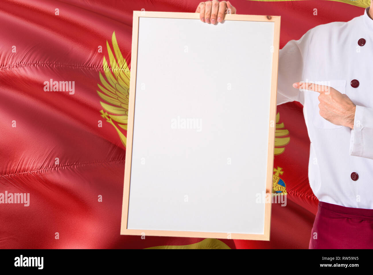 Montenegrin Chef holding blank whiteboard menu on Montenegro flag background. Cook wearing uniform pointing space for text. Stock Photo