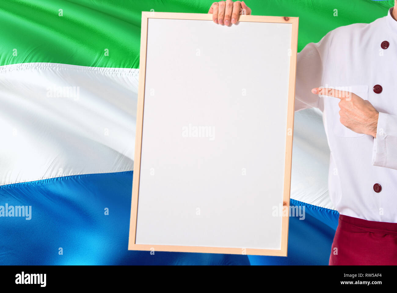 Chef holding blank whiteboard menu on Sierra Leone flag background. Cook wearing uniform pointing space for text. Stock Photo