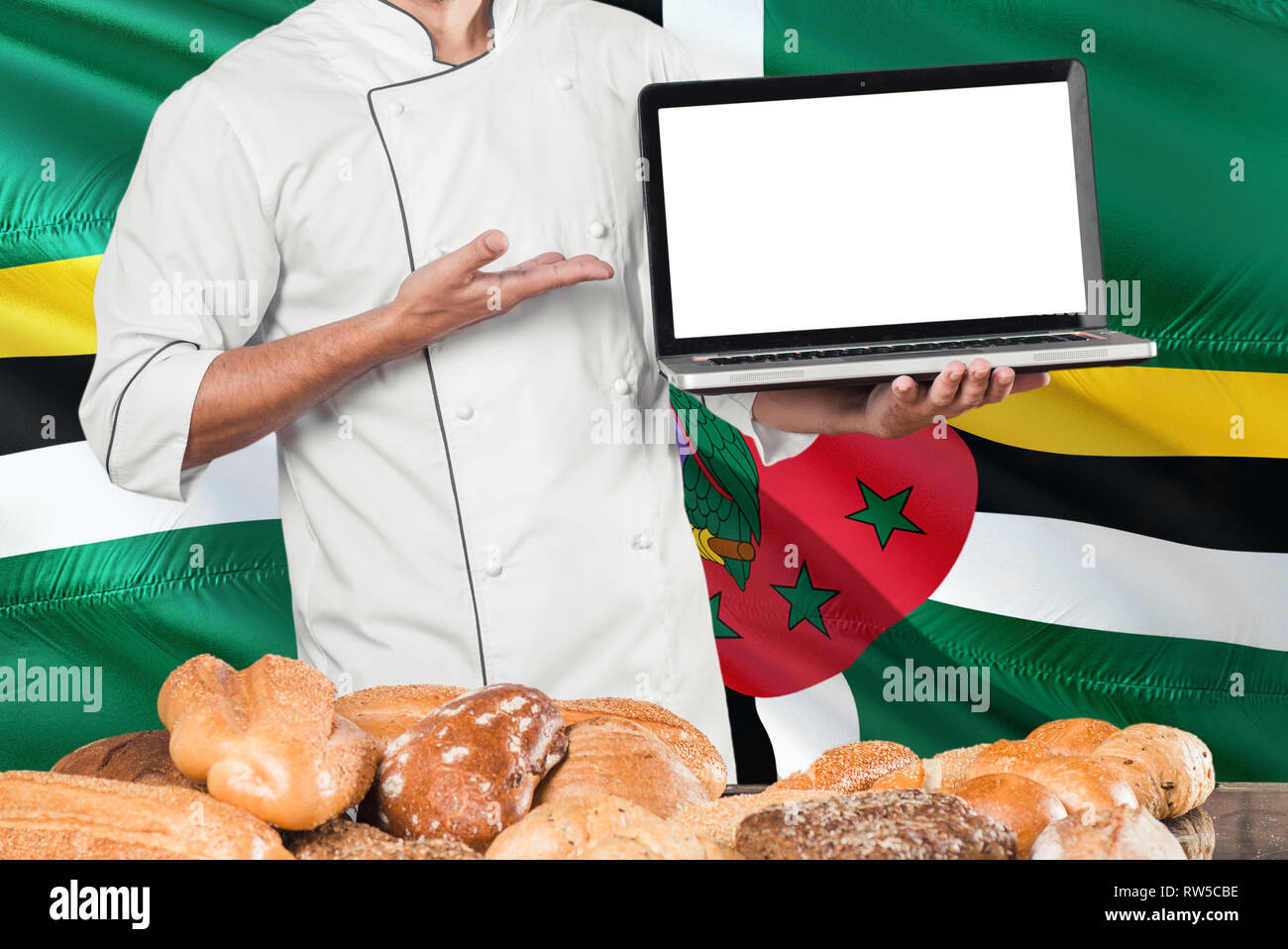 Dominican Baker holding laptop on Dominica flag and breads background. Chef wearing uniform pointing blank screen for copy space. Stock Photo