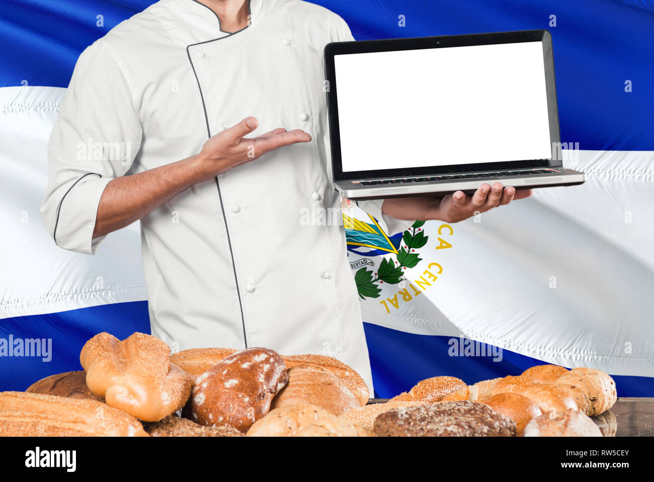 Salvadoran Baker holding laptop on El Salvador flag and breads background. Chef wearing uniform pointing blank screen for copy space. Stock Photo