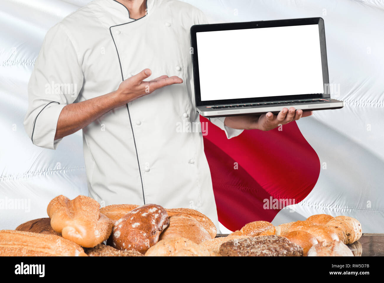 Japanese Baker holding laptop on Japan flag and breads background. Chef wearing uniform pointing blank screen for copy space. Stock Photo