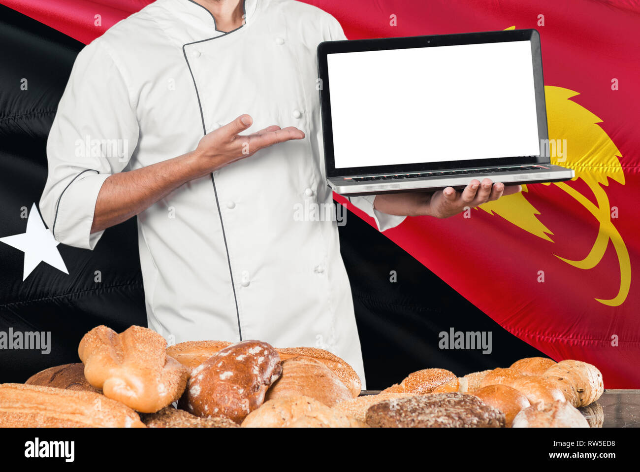 Baker holding laptop on Papua New Guinea flag and breads background. Chef wearing uniform pointing blank screen for copy space. Stock Photo