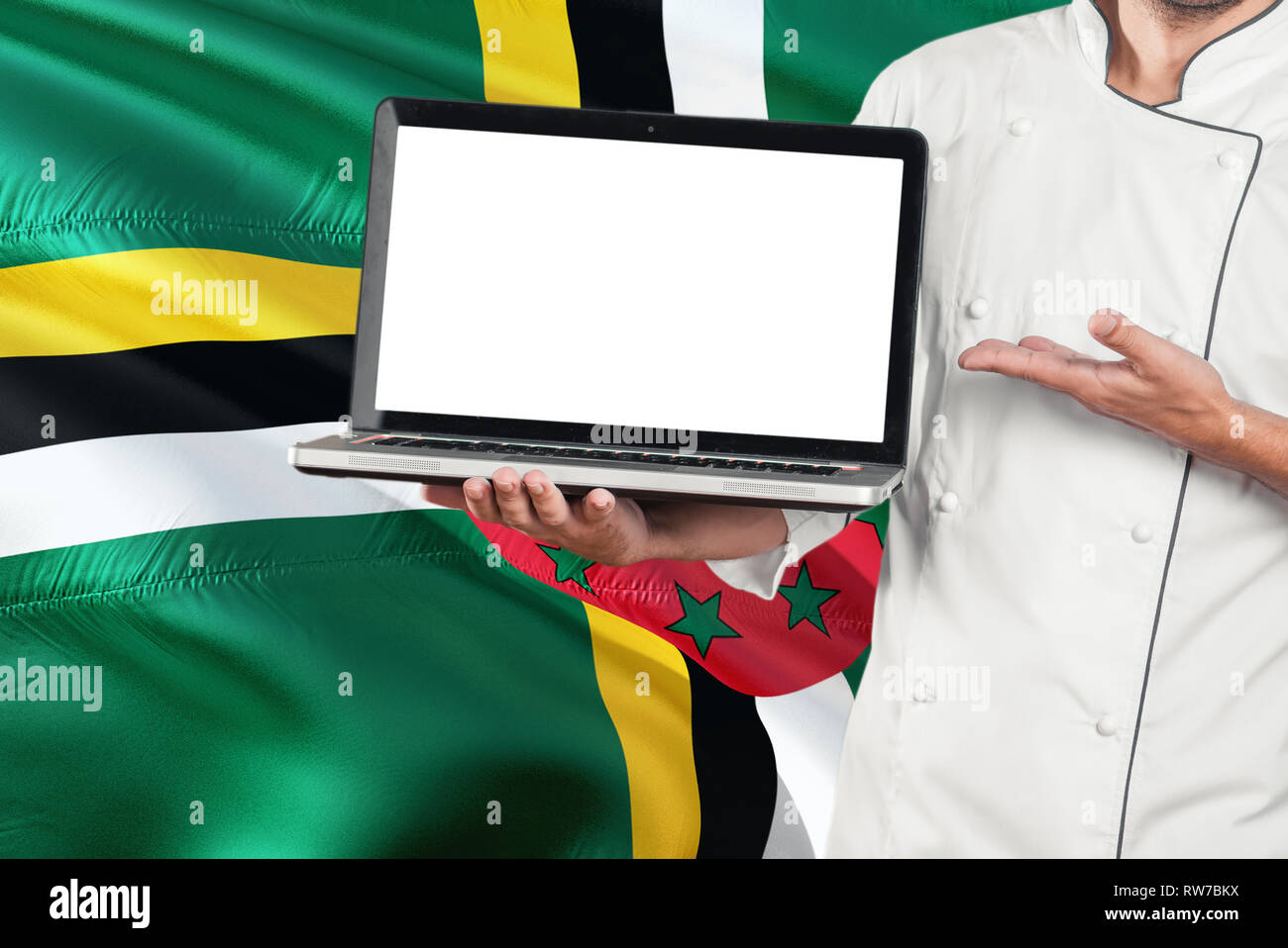 Dominican Chef holding laptop with blank screen on Dominica flag background. Cook wearing uniform and pointing laptop for copy space. Stock Photo