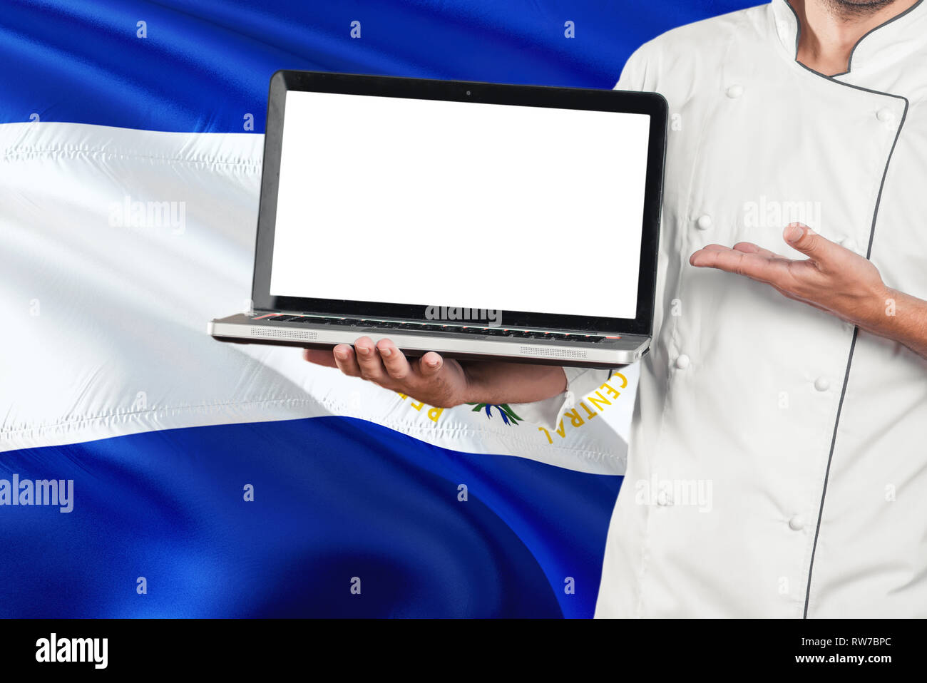 Salvadoran Chef holding laptop with blank screen on El Salvador flag background. Cook wearing uniform and pointing laptop for copy space. Stock Photo