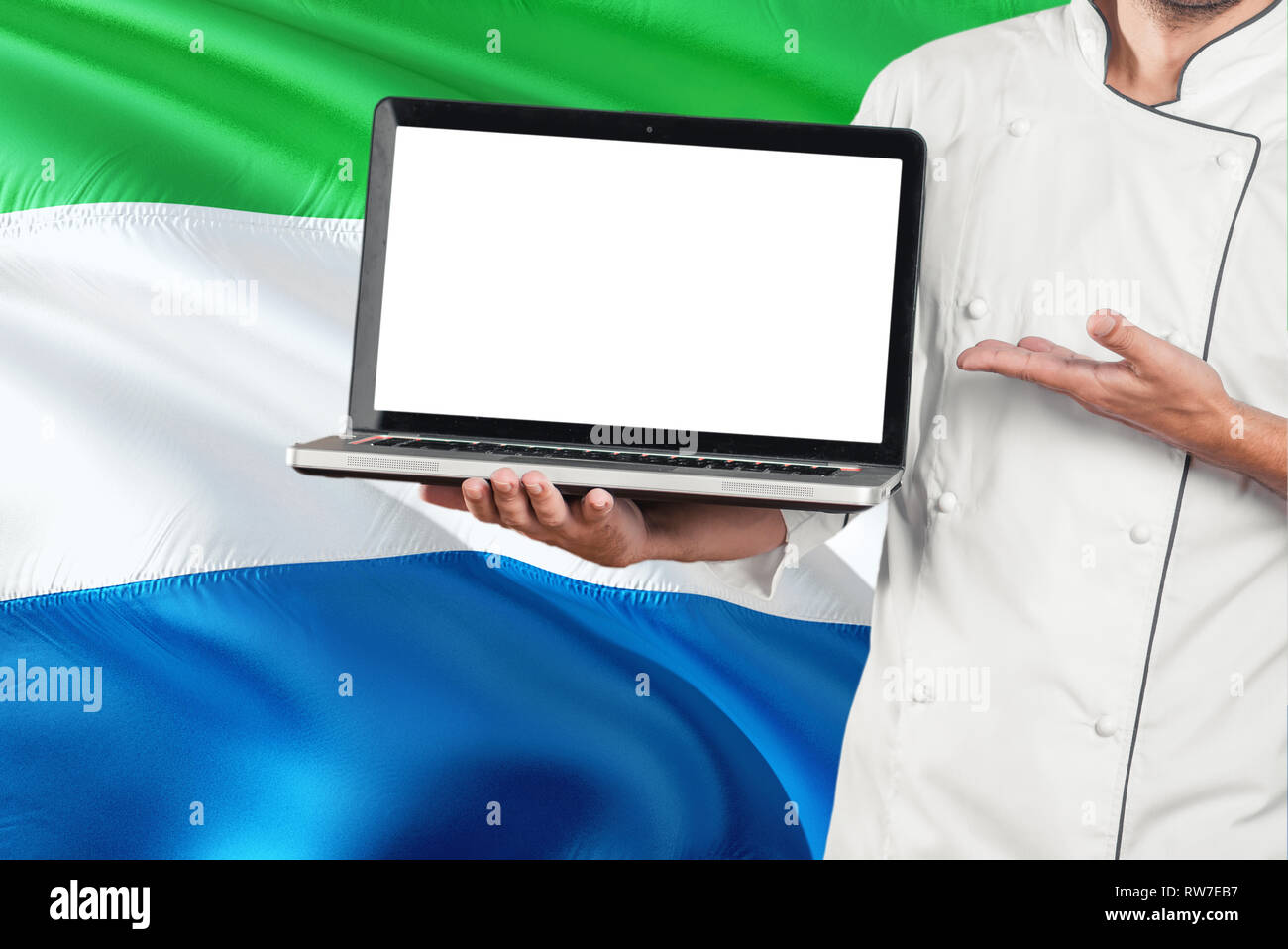 Chef holding laptop with blank screen on Sierra Leone flag background. Cook wearing uniform and pointing laptop for copy space. Stock Photo