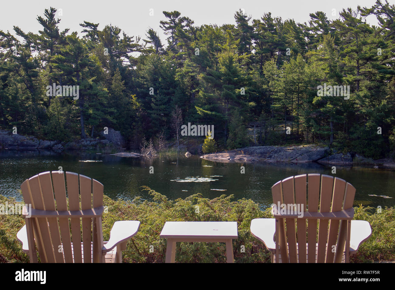 Two Muskoka chairs sitting on a wood dock facing a calm lake. Across ...