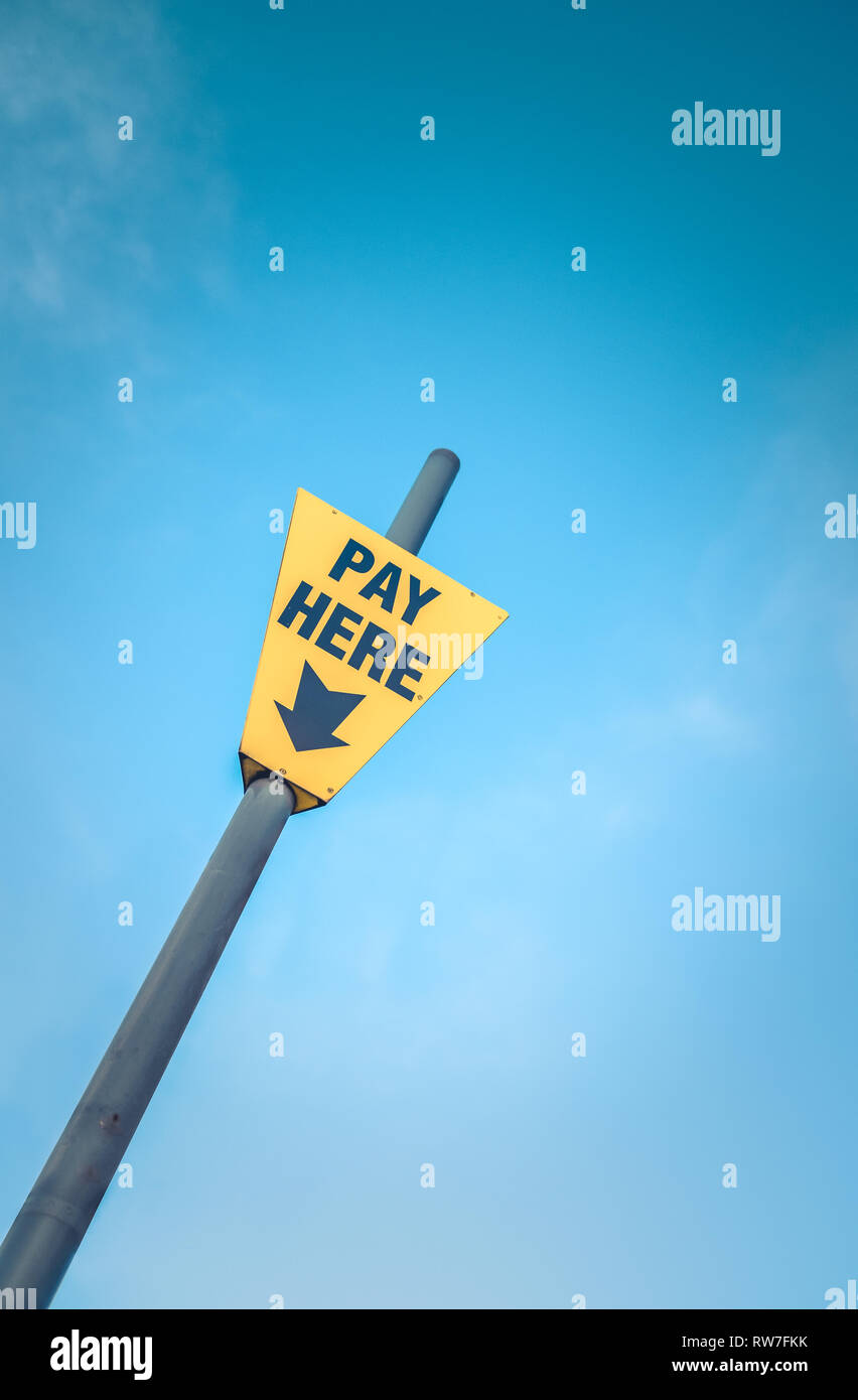 Conceptual Image Of A Bright Yellow Pay Here Sign Against A Blue Sky With Copy Space Stock Photo