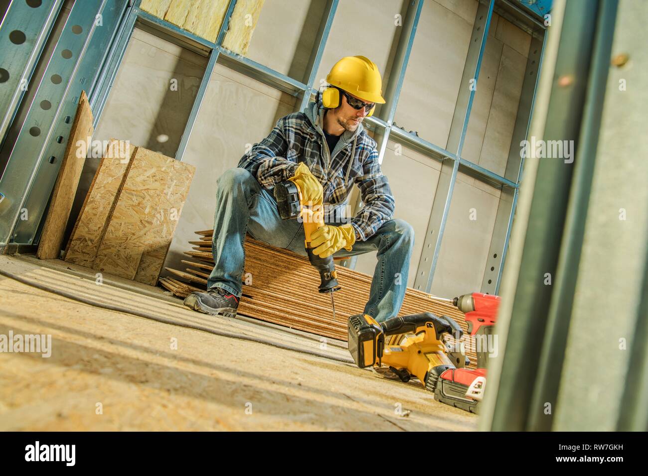 Construction Worker with Cordless Reciprocating Saw in Hands Taking Short Brake While Seats on the Pile of Plywood Boards. Construction Industry Theme Stock Photo