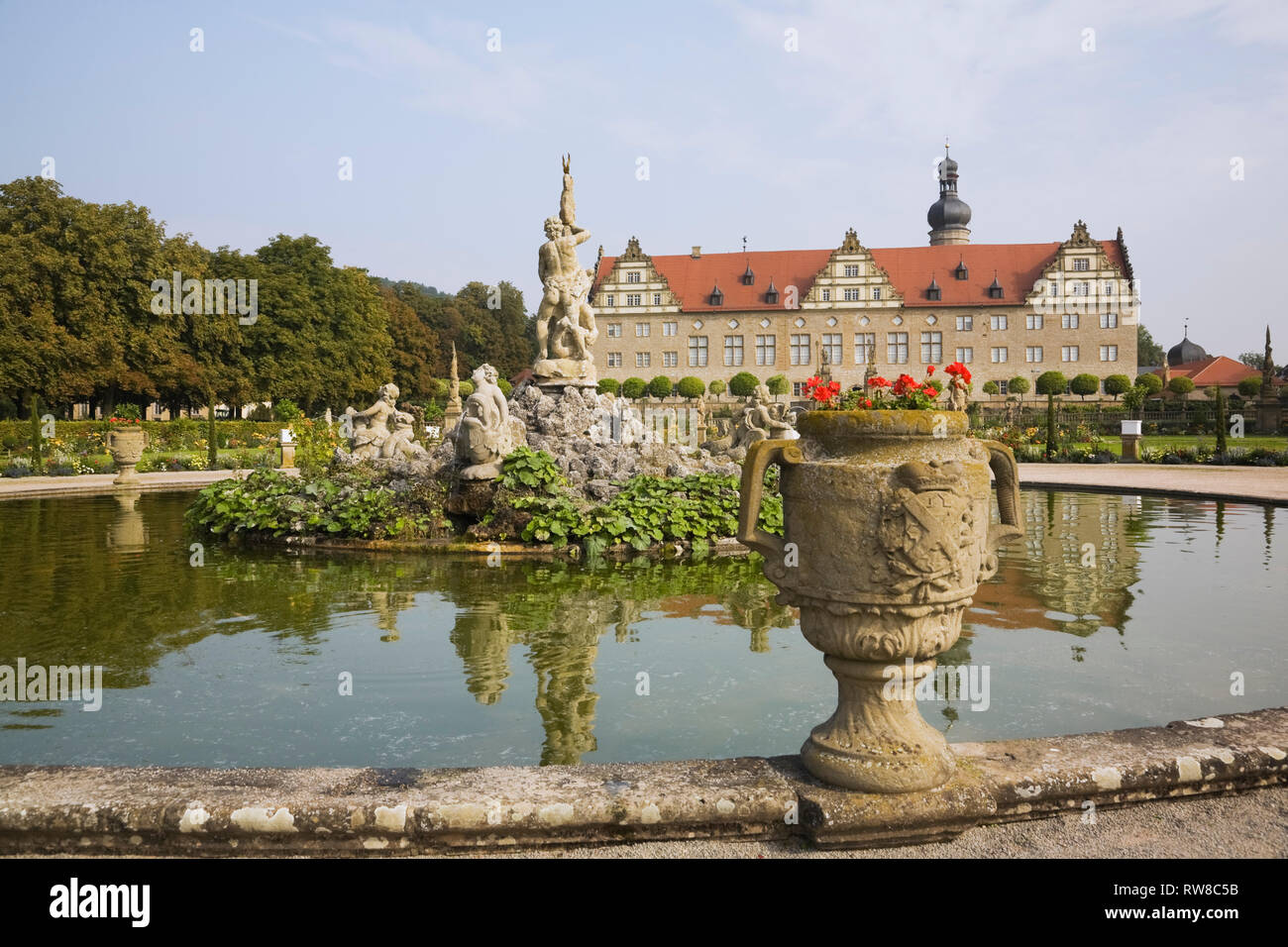 Red Geranium flowers in planter and water fountain pool with statue at the Weikersheim Palace garden in late summer, Hohenlohe region, Germany Stock Photo